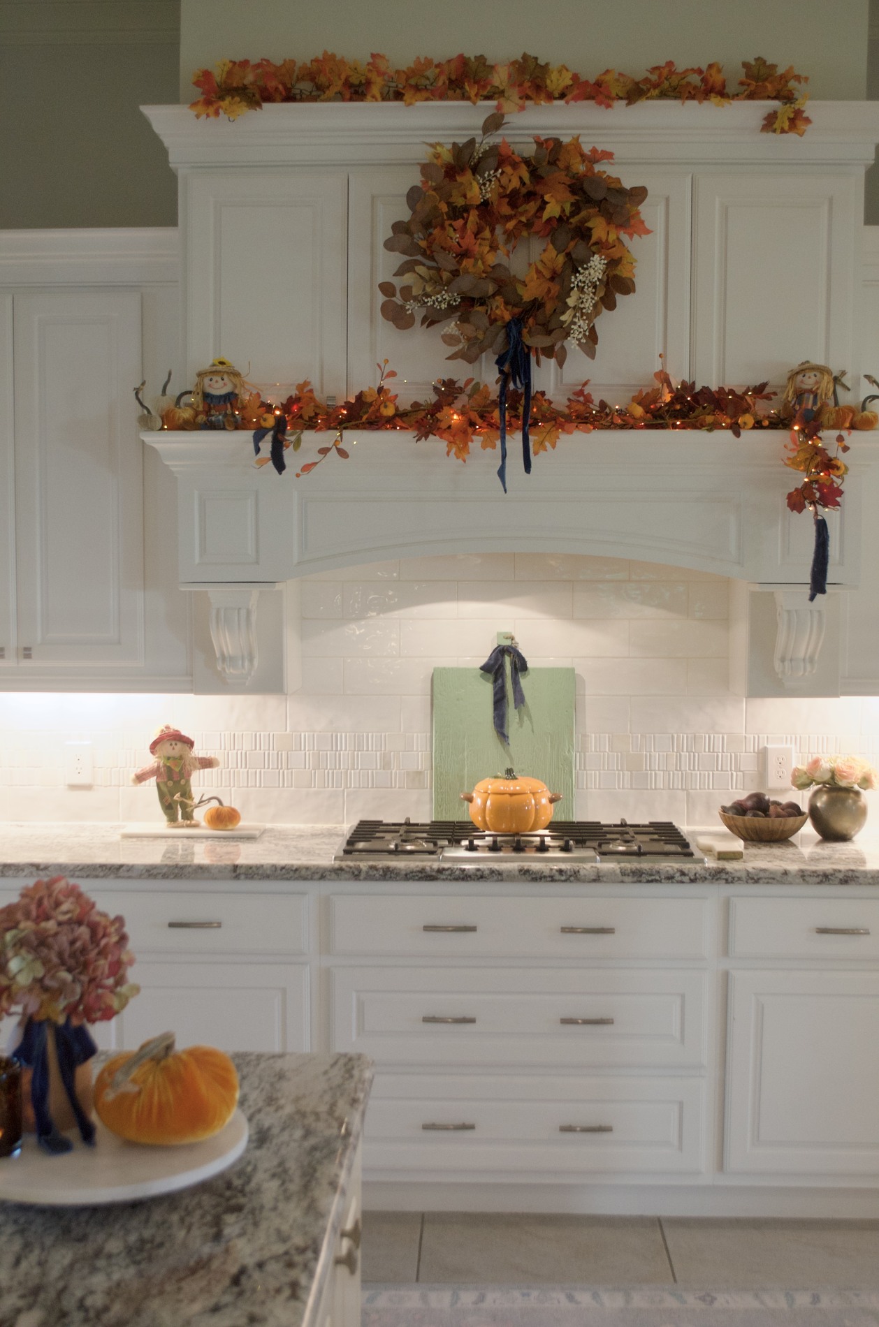 White kitchen decorated with an autumn wreath, orange and golden leafy garlands, small scarecrows, pumpkins, blue ribbon, and an orange pumpkin-shaped pot on the cooktop.