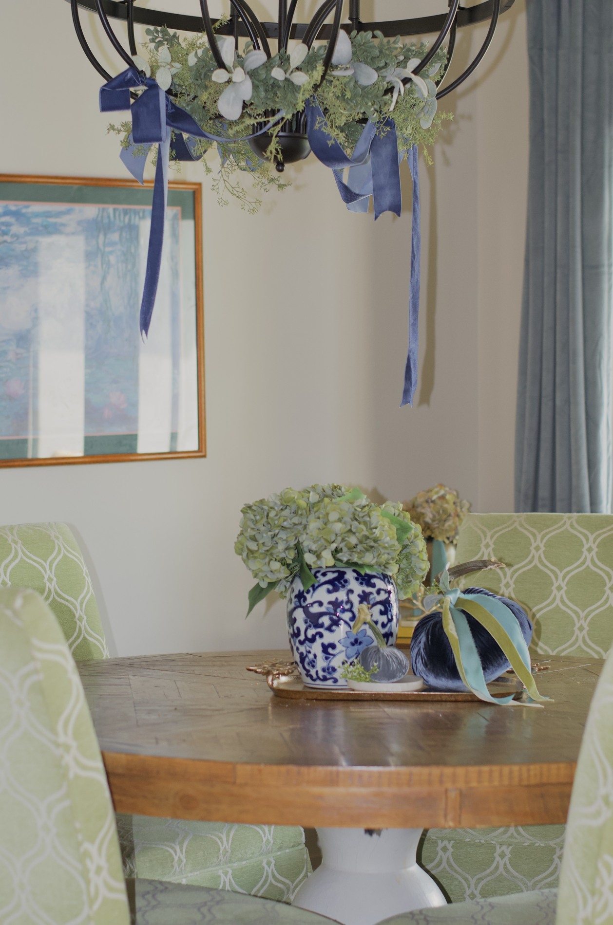 Breakfast room centerpiece with hydrangeas, a blue-and-white vase, navy and blue-gray pumpkins, and blue and green ribbon beneath a chandelier wrapped in greenery and blue ribbon.
