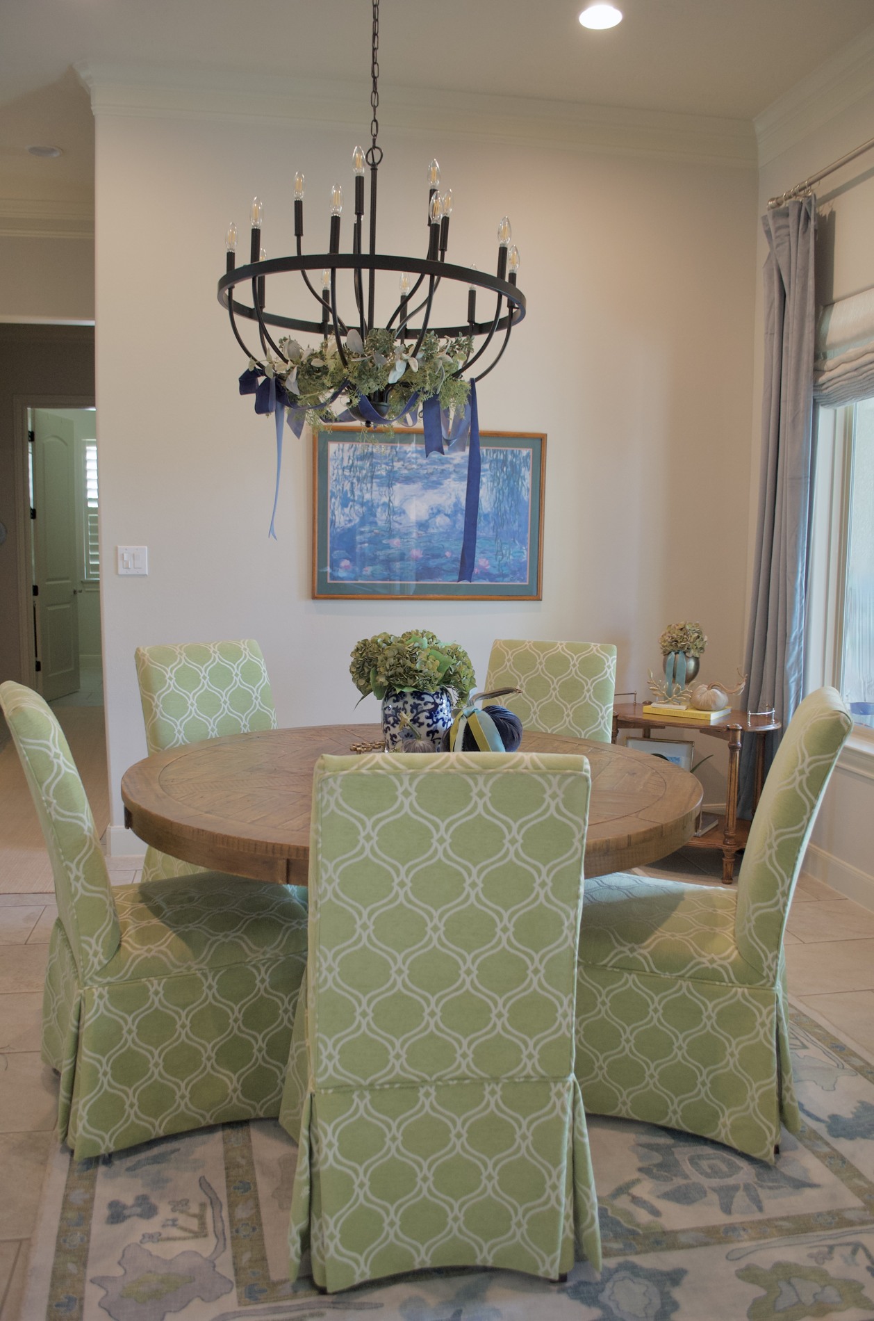 Round wooden breakfast table with green patterned chairs, hydrangeas in a blue-and-white vase, velvet pumpkins, and a chandelier decorated with greenery and blue ribbon.