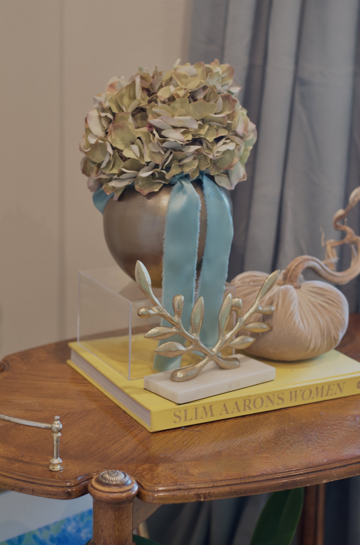 Breakfast room side-table close-up with hydrangeas in a metallic vase, long blue ribbon, an ivory velvet pumpkin, gold branch sculpture, and stacked books.