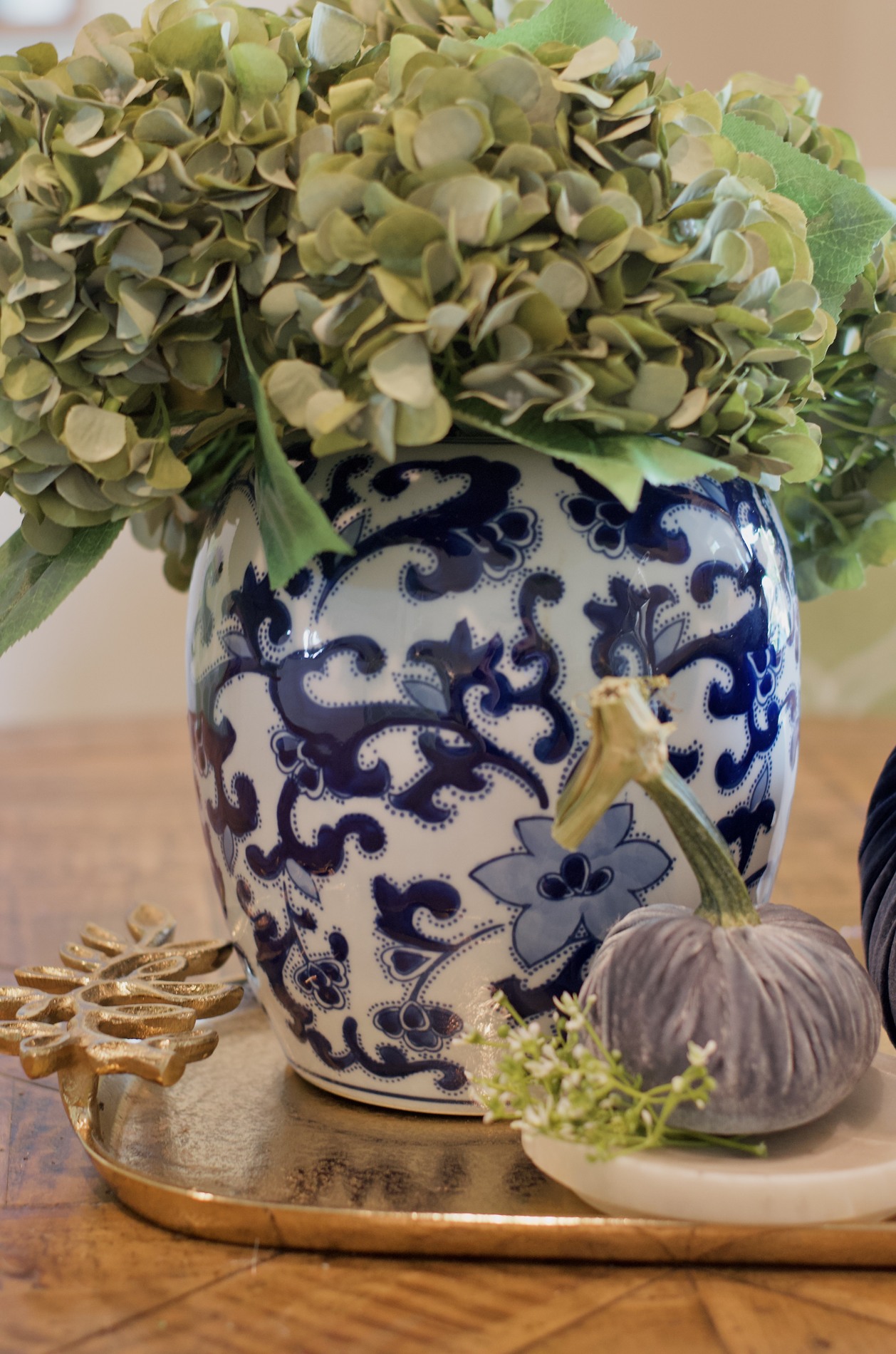 Close-up of green hydrangeas in a blue-and-white vase beside a small blue-gray velvet pumpkin and a gold leafy tray on the breakfast table.