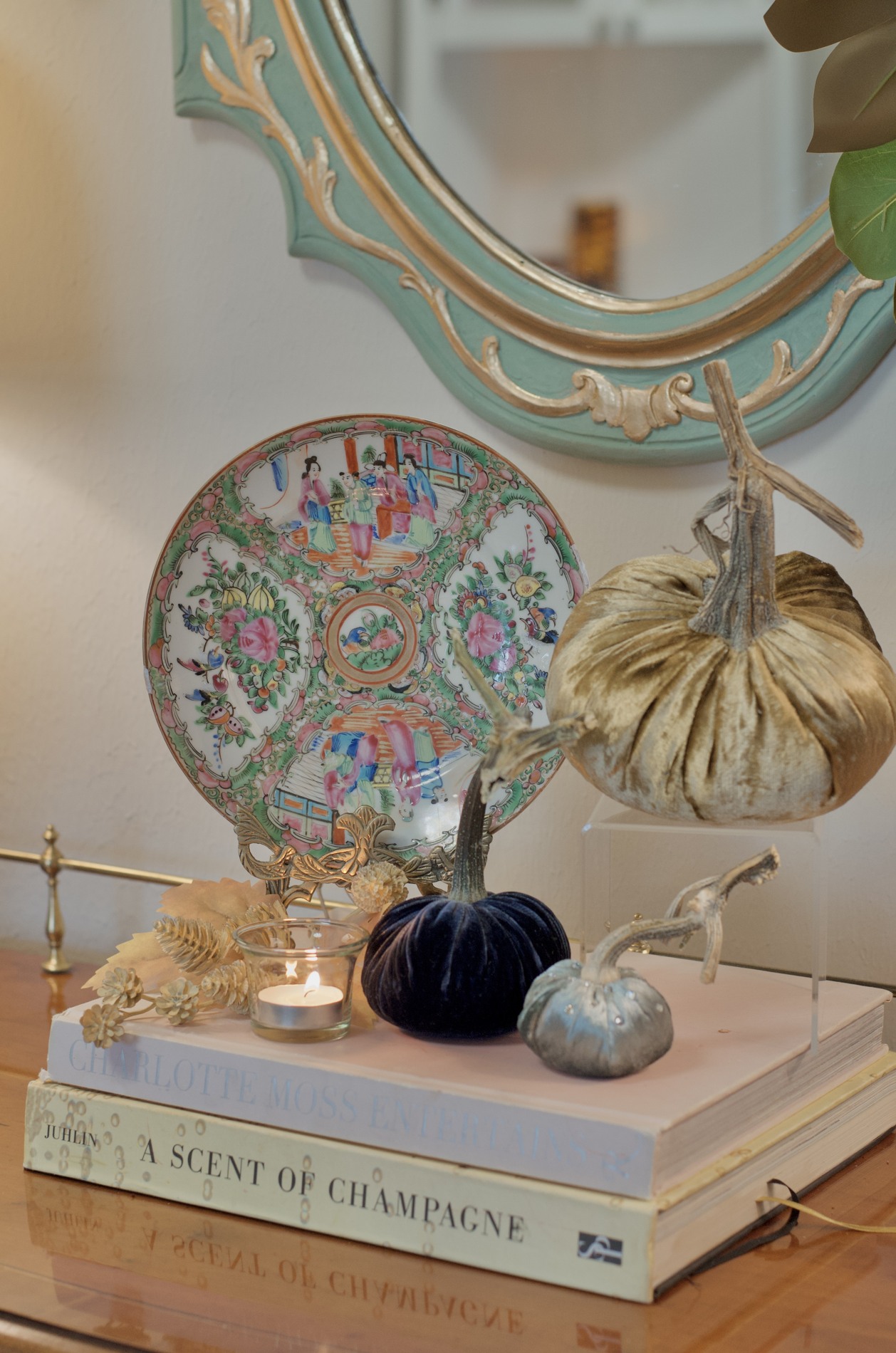 Close-up of a colorful decorative plate beside gold, navy, and pale blue velvet pumpkins on stacked books and a clear riser beneath a turquoise-and-gold mirror.