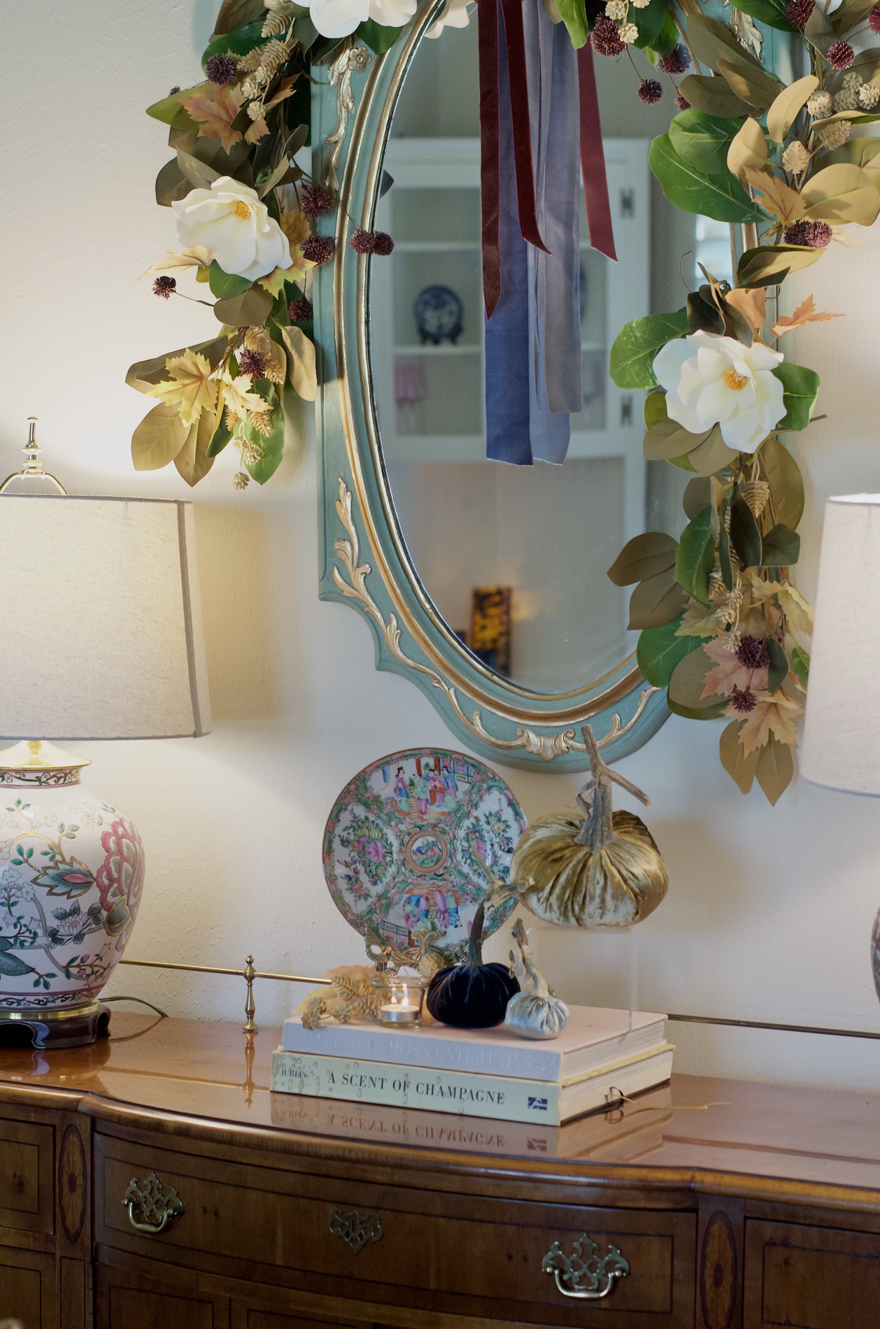 Turquoise-and-gold dining room mirror framed with magnolia greenery, white blooms, golden foliage, and burgundy and blue ribbon above a decorated wood buffet.