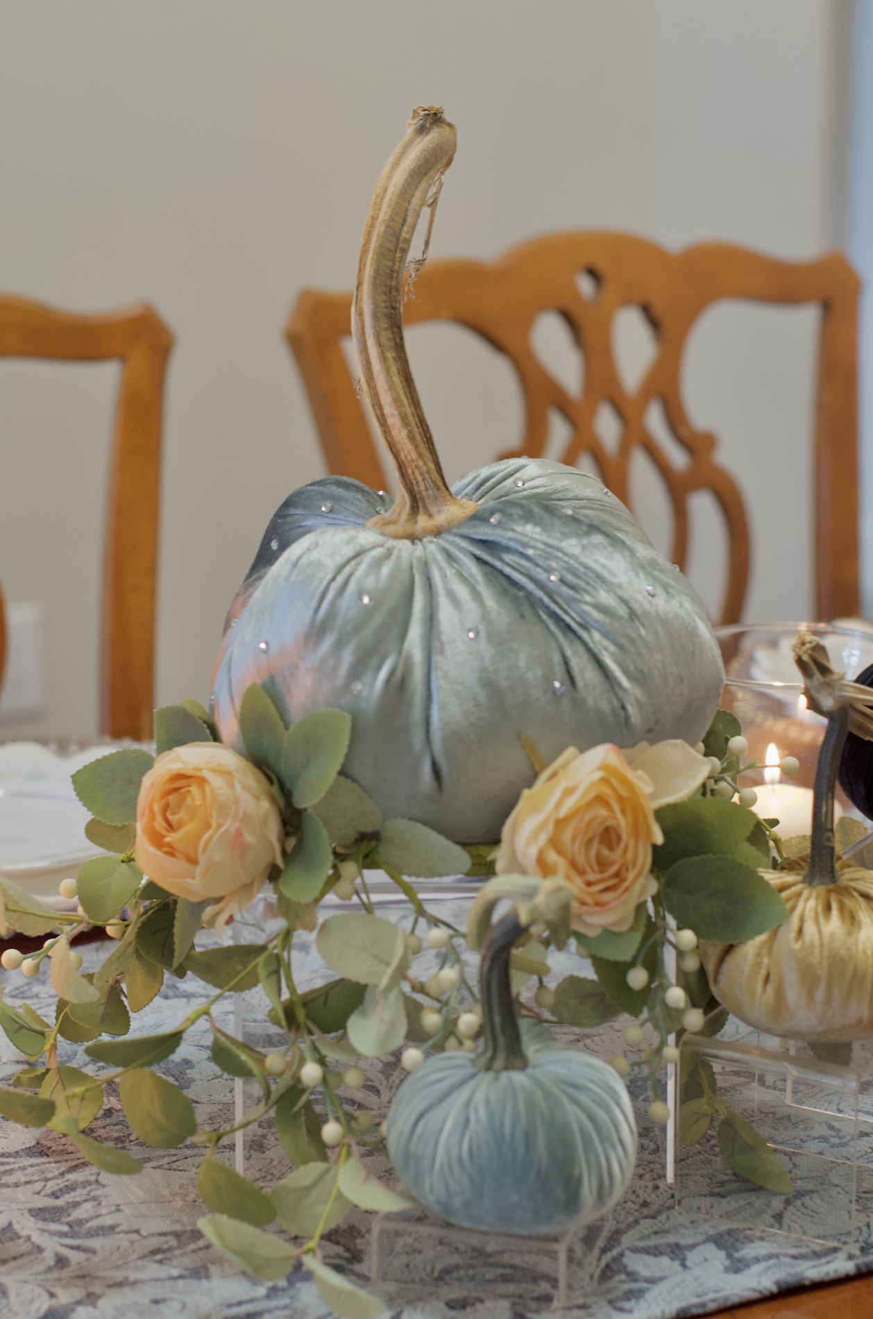 Close-up of an embellished pale blue velvet pumpkin with a tall natural stem, surrounded by cream roses, greenery, smaller pumpkins, and a glass candleholder.