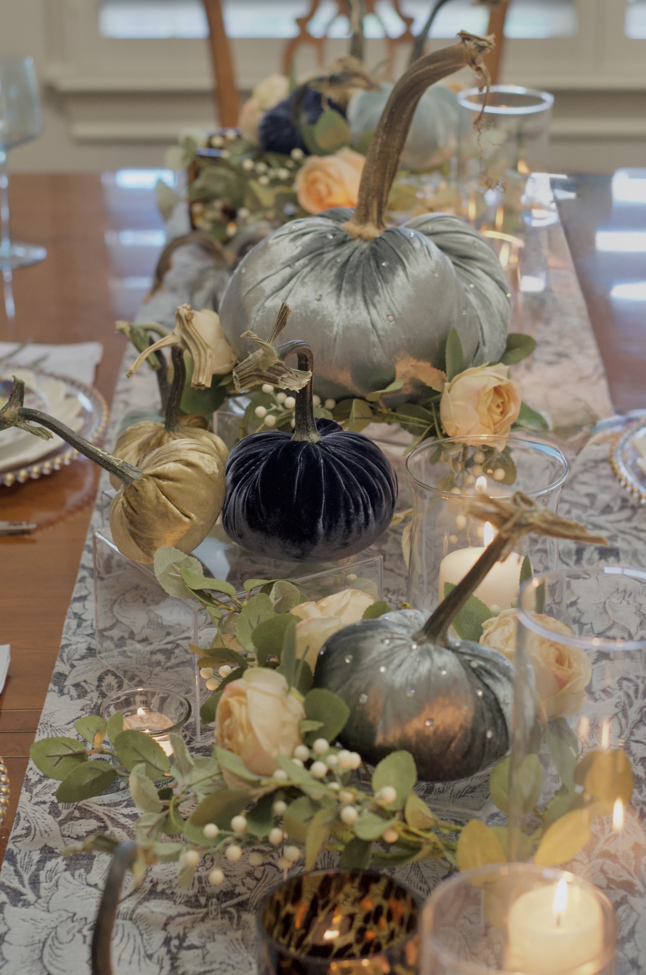 Close-up along a dining centerpiece with pale blue, navy, and gold velvet pumpkins at different heights, cream roses, greenery, and glass candleholders.