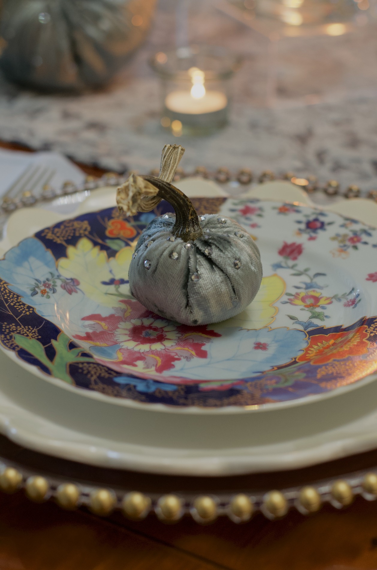 Tight close-up of an embellished miniature blue-gray velvet pumpkin with a natural stem on a colorful floral plate, layered over a white plate and a gold-edged charger.