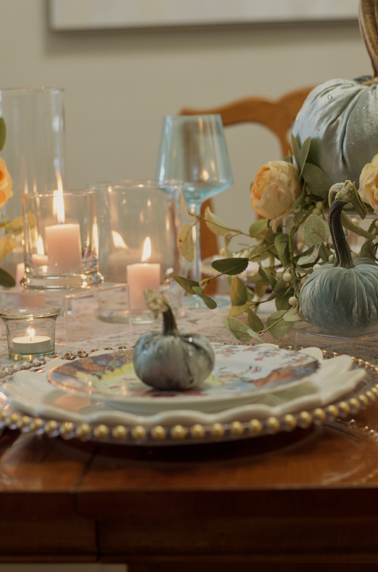Close-up of a miniature blue-gray pumpkin on a colorful plate over scalloped white layers, with pale blue stemware, candlelight, and a velvet pumpkin centerpiece behind.
