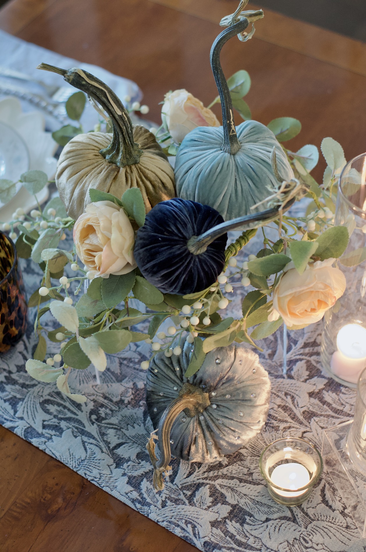 Overhead close-up of gold, blue-green, navy, and embellished blue-gray velvet pumpkins among cream roses, green leaves, pale berries, and a glass candleholder.
