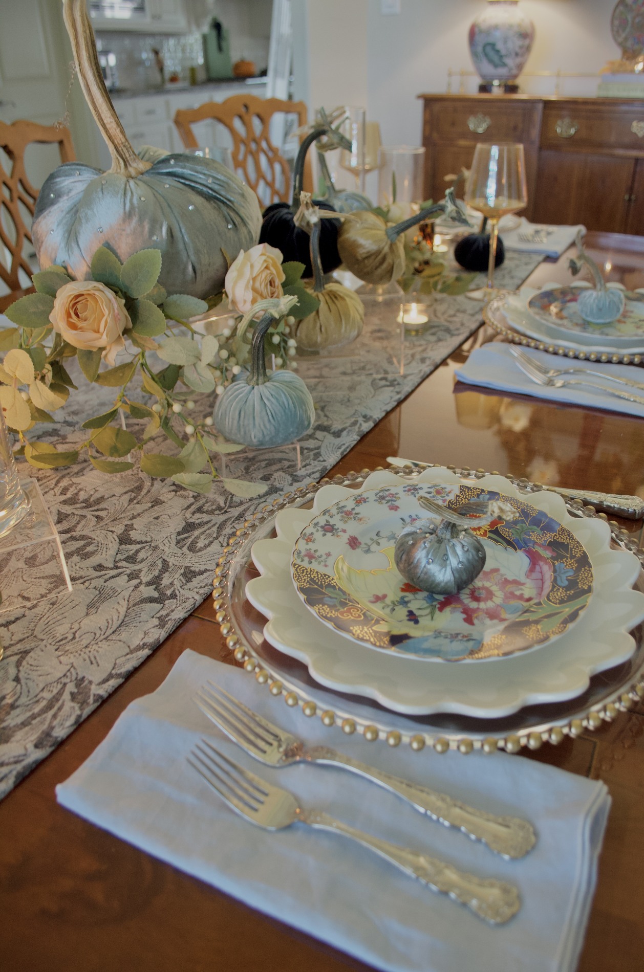 Dining table with a blue-gray runner, velvet pumpkins on clear risers, cream roses, greenery, glass candleholders, and layered colorful place settings.