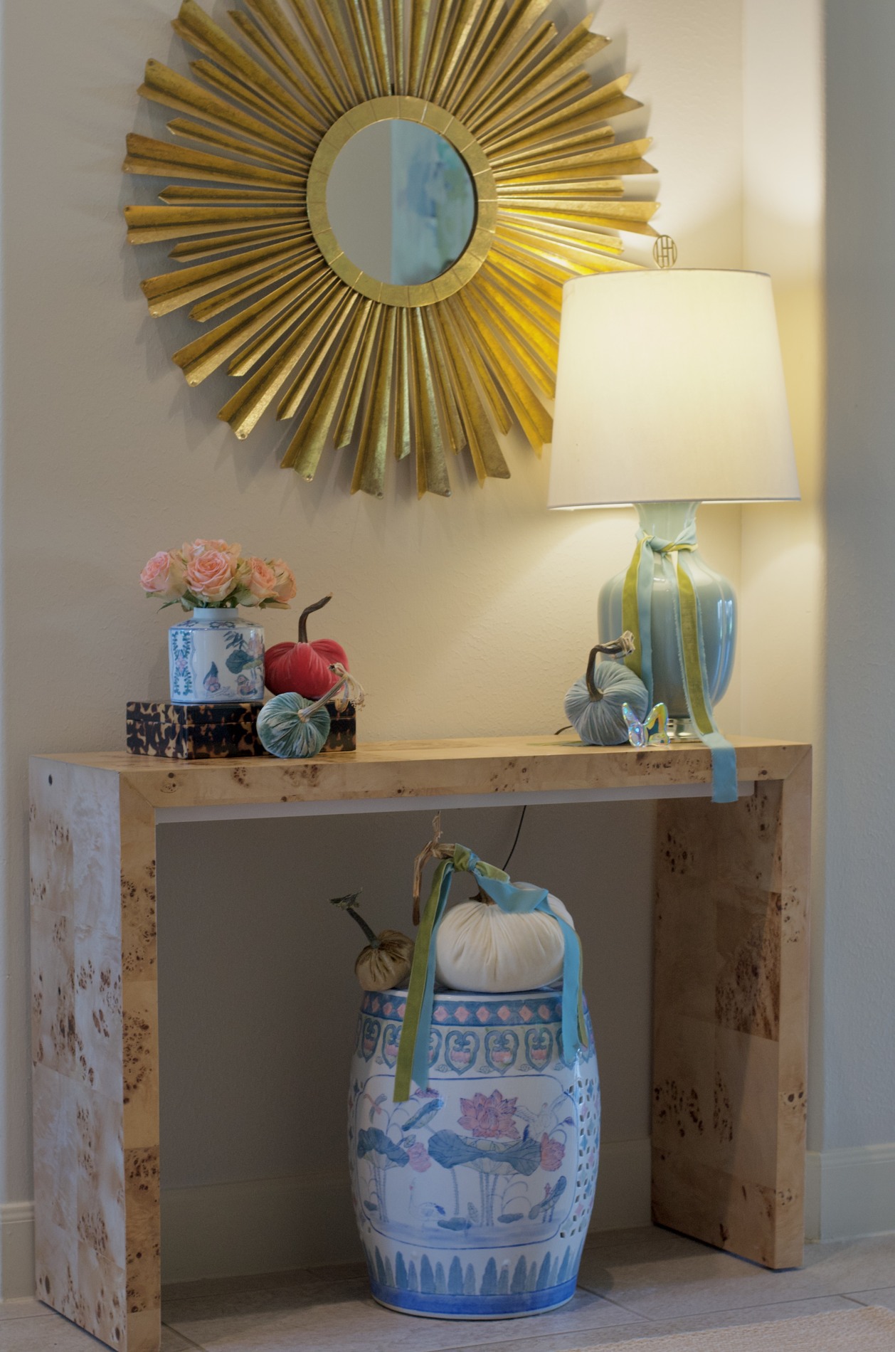 Wood foyer console beneath a gold sunburst mirror, with a pale blue lamp, floral ceramic vase, red and blue-green pumpkins, and an ivory pumpkin on a patterned garden stool below.