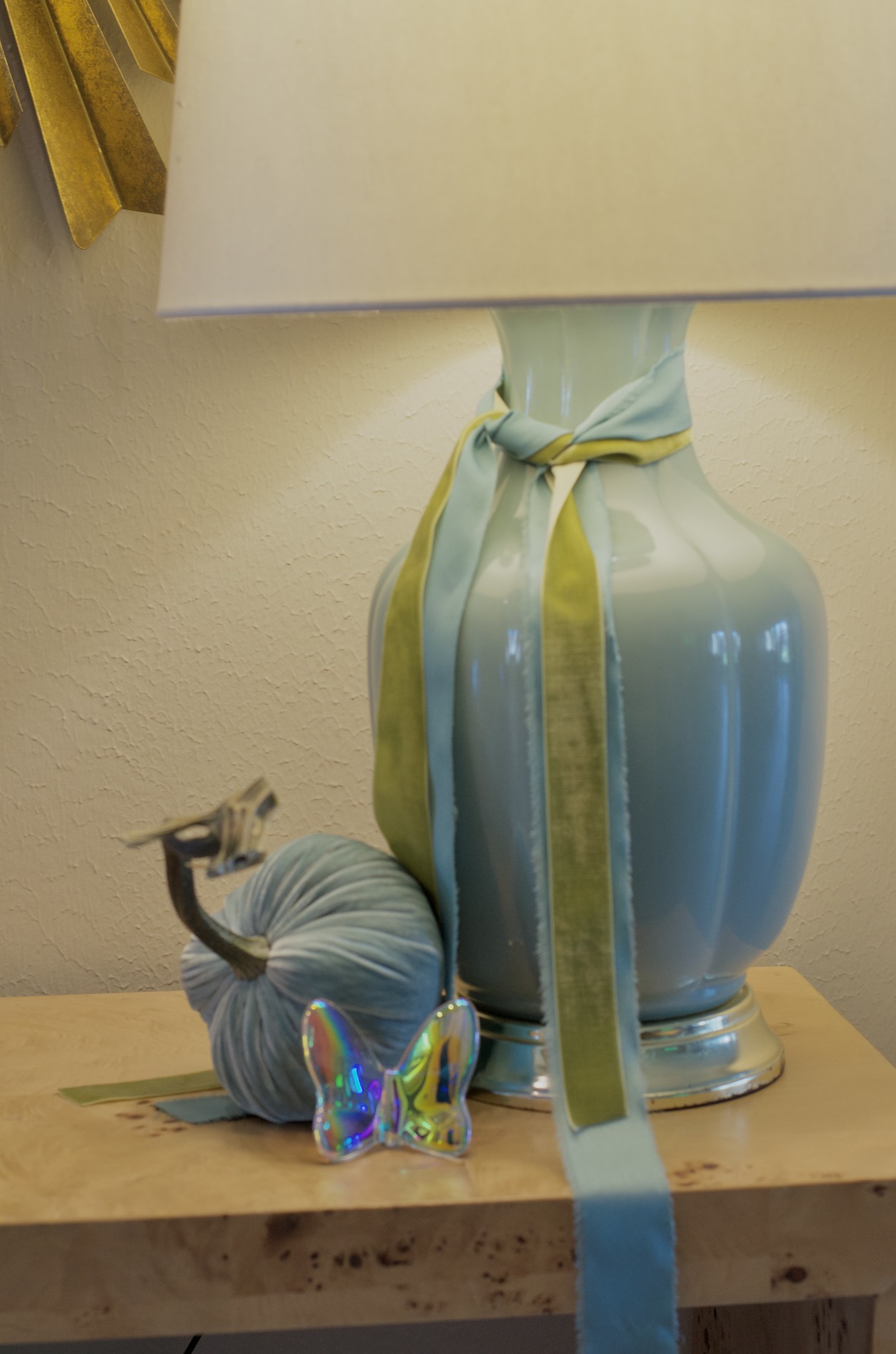 Close-up of a pale blue foyer lamp tied with blue and green ribbon, a blue-gray velvet pumpkin, and an iridescent glass butterfly on a wood console.
