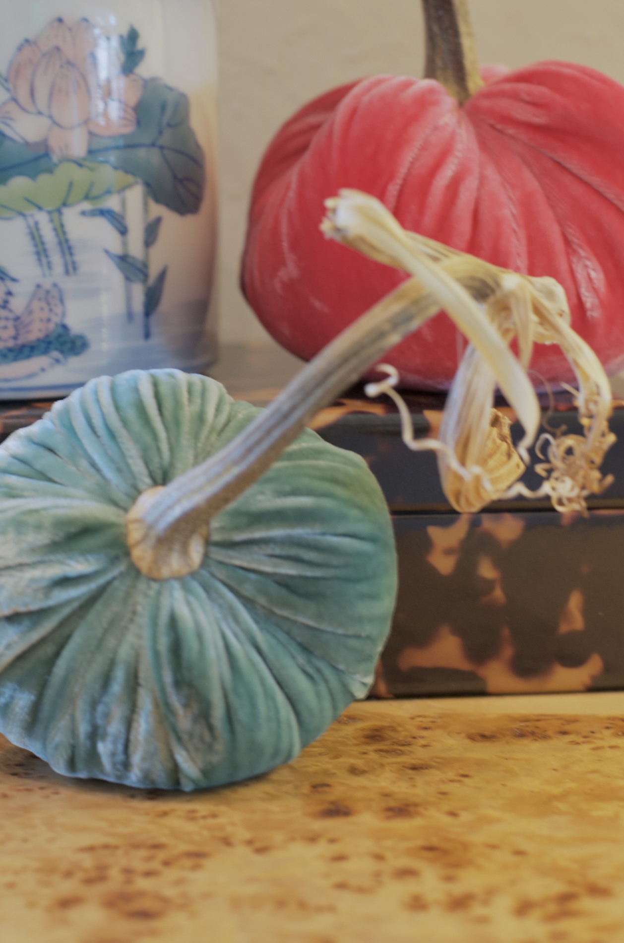 Close-up of red and blue-green velvet pumpkins with natural stems beside a floral ceramic vase and patterned box on the foyer table.