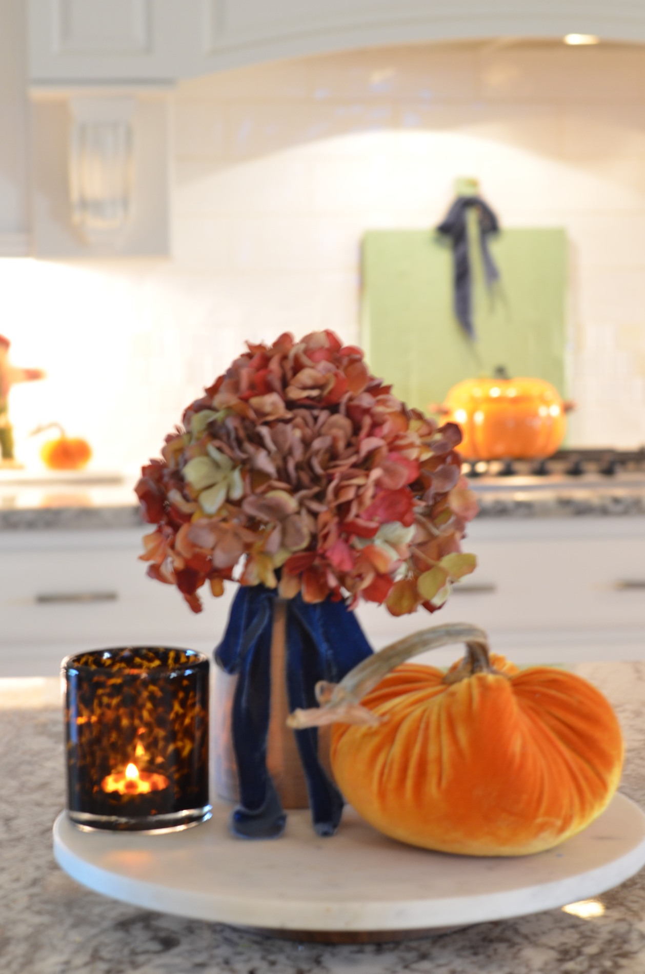 Kitchen island close-up of an orange velvet pumpkin with a real stem, rust and pink-toned hydrangeas with navy ribbon, and a lit amber patterned candle arranged on a round pale tray.