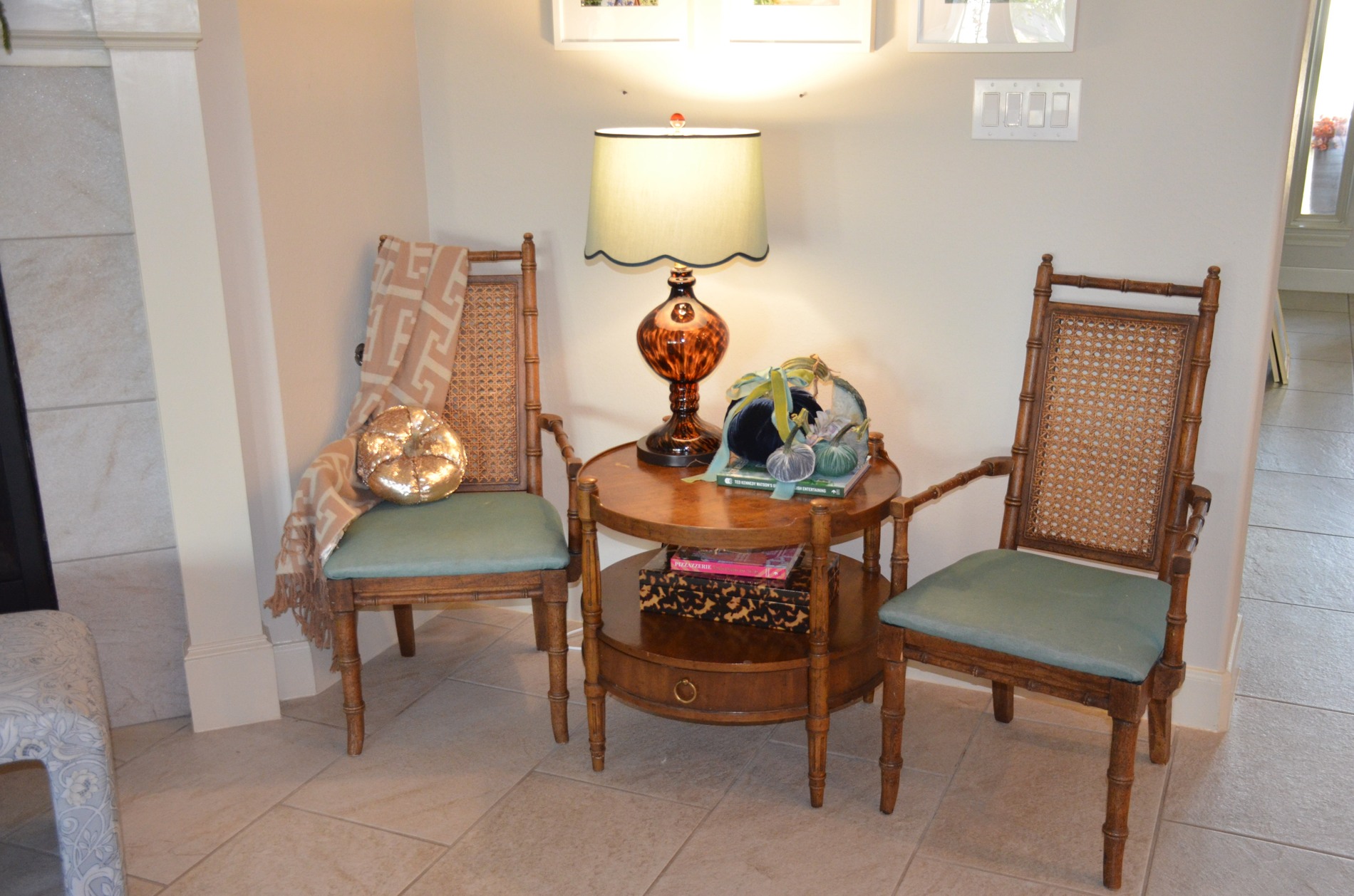Round wooden side table between two bamboo and cane chairs with green cushions, decorated with a patterned amber lamp, navy and smaller blue-gray and green velvet pumpkins, blue and green ribbon, and books.