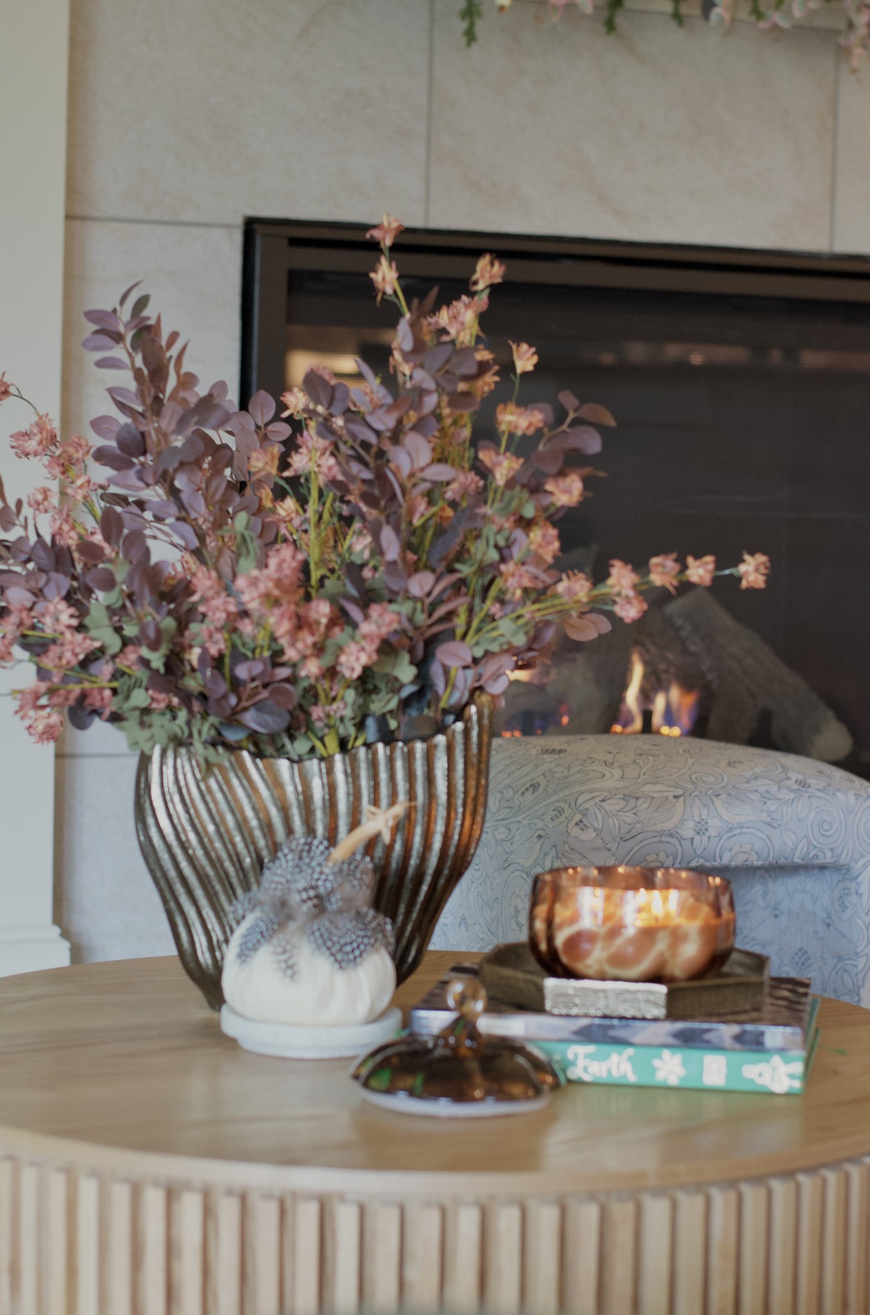 Coffee table close-up with burgundy and pink foliage in a ribbed metallic vase, a small embellished ivory pumpkin, stacked books, and a lit amber patterned candle beside the fireplace.