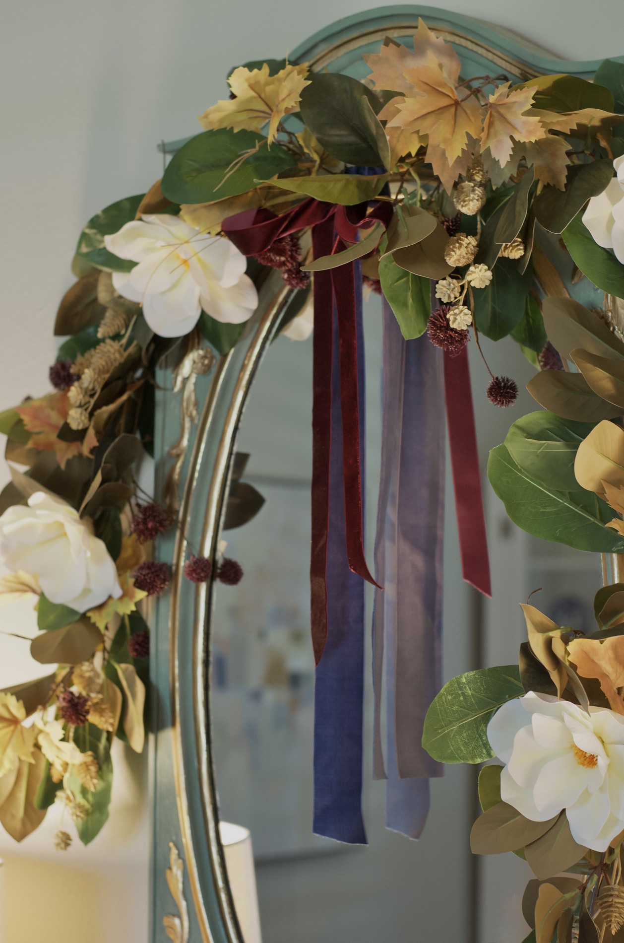 Close-up of magnolia leaves and white blooms with golden autumn leaves, small cream and burgundy accents, and burgundy ribbon layered with blue ribbon on a mirror garland.
