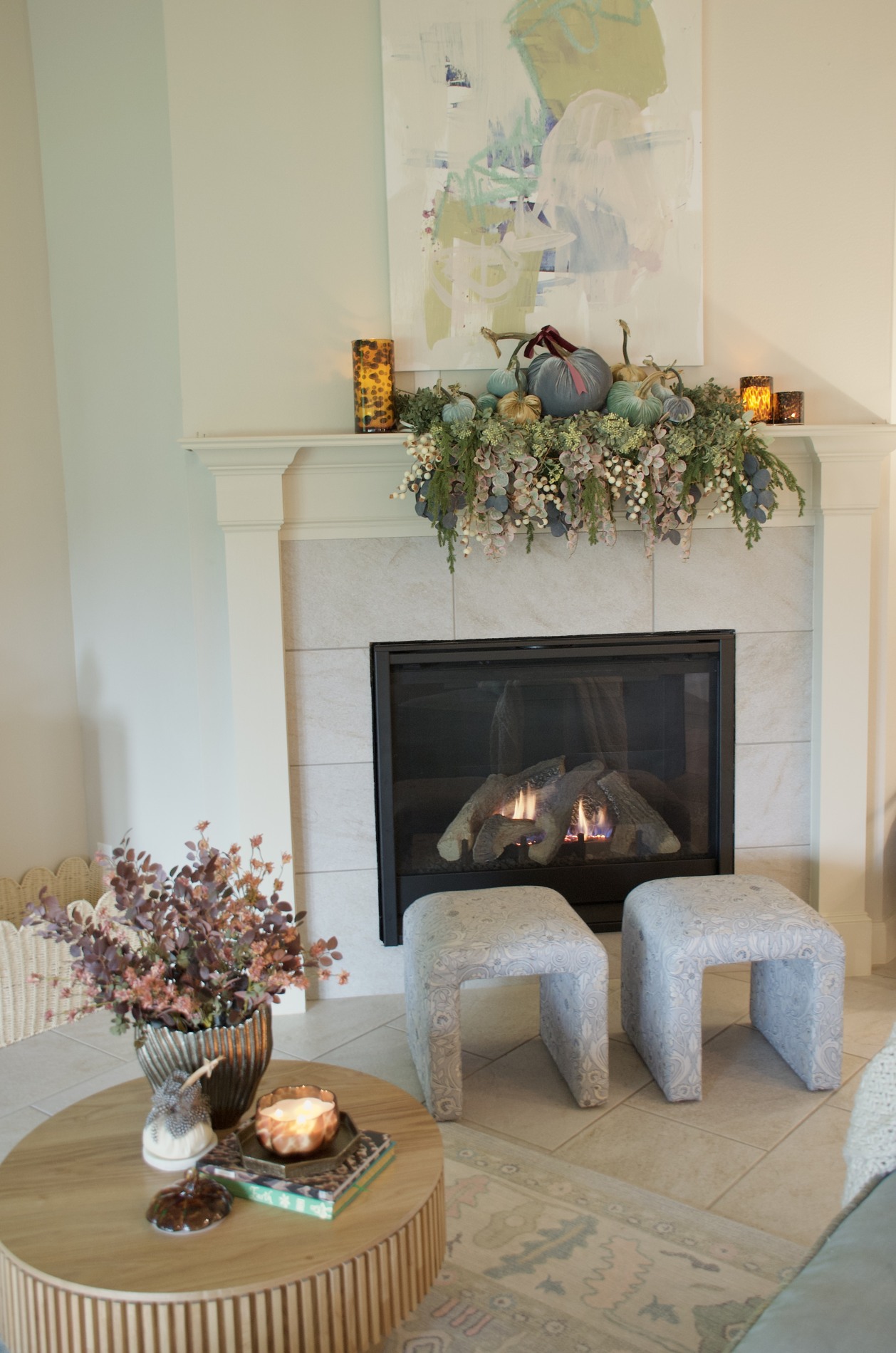 White fireplace beneath abstract artwork, decorated with blue, green, and gold velvet pumpkins, burgundy ribbon, trailing greenery, and warm lights.