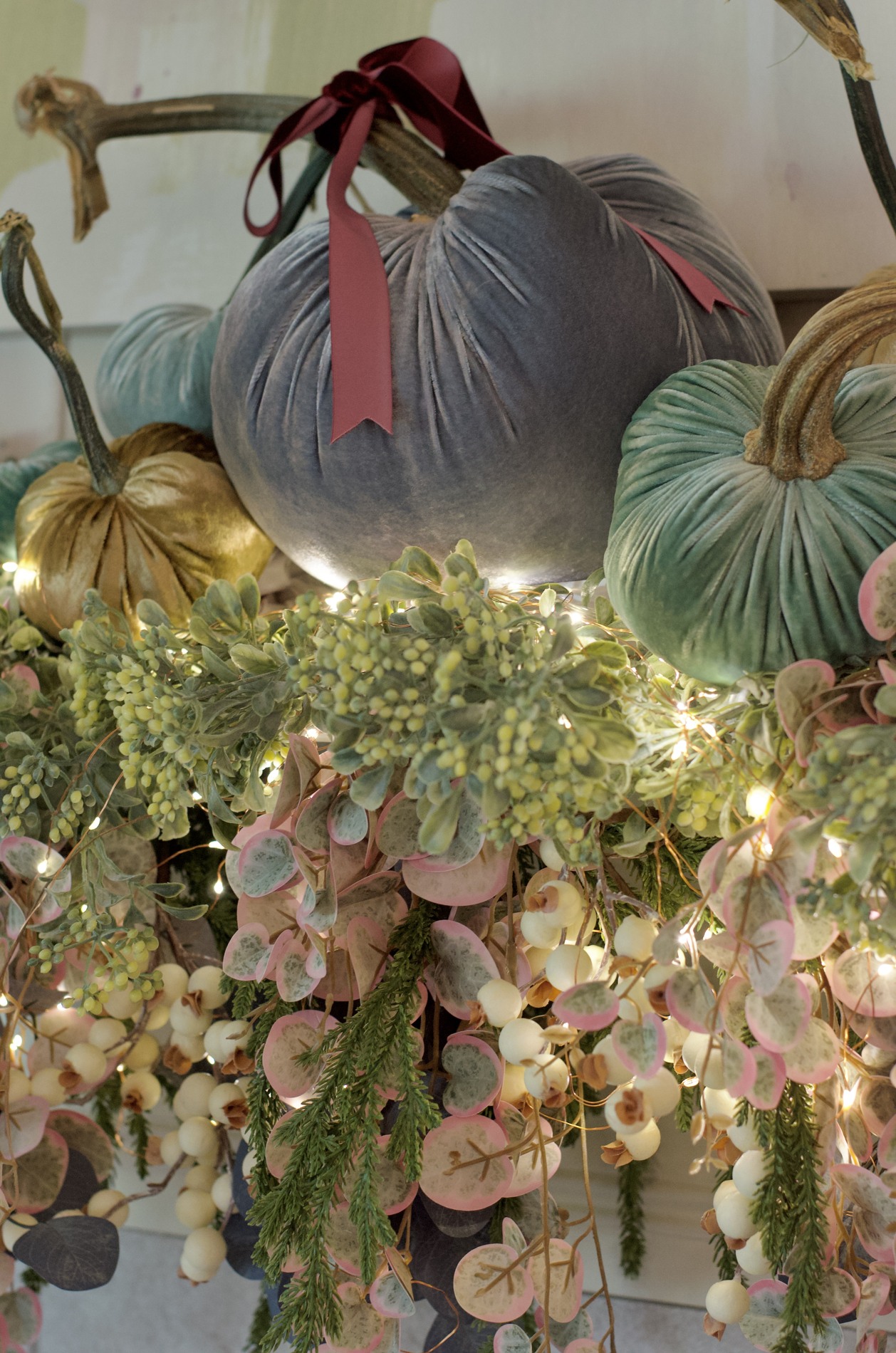 Close-up of illuminated mantel garland with pale berries, green and pink-edged leaves, blue-gray and green velvet pumpkins, and burgundy ribbon.