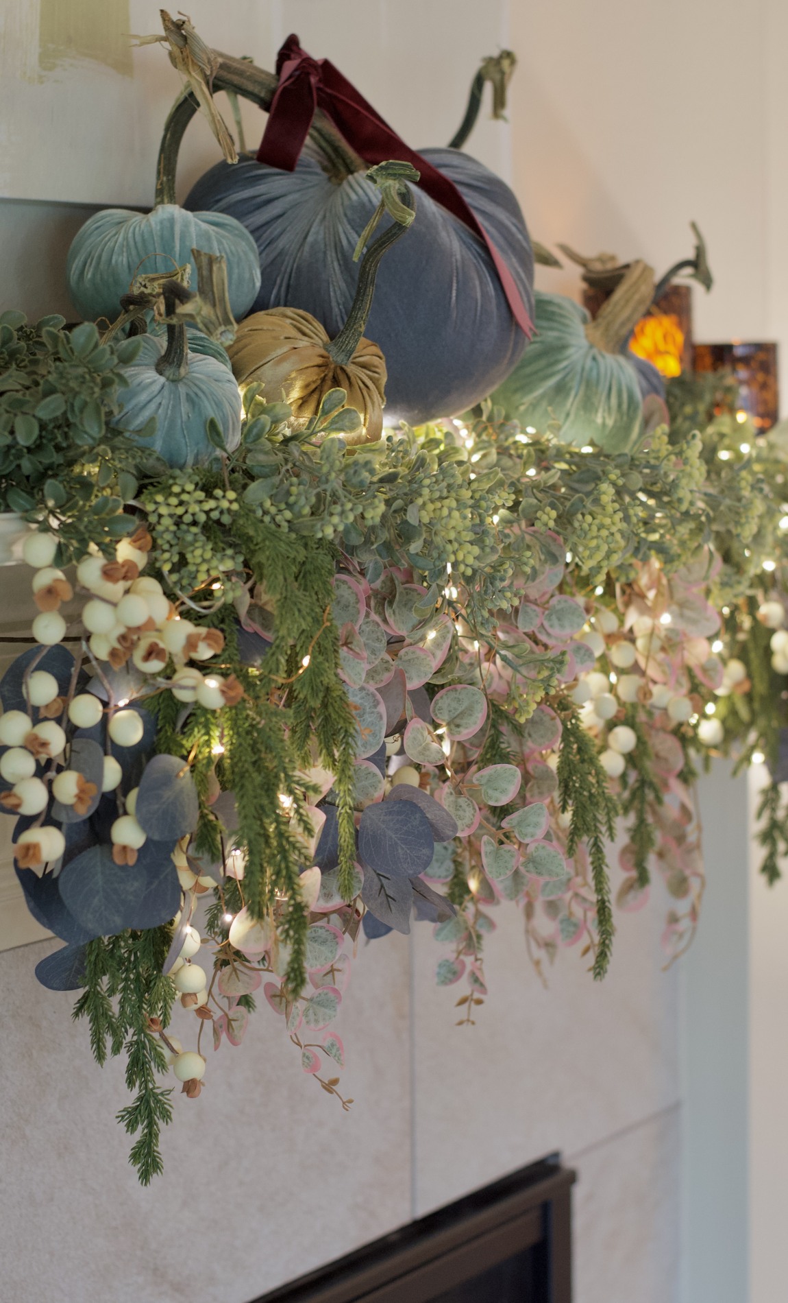 Angled close-up of lit mantel garland with trailing green, blue-gray, and pink-edged foliage beneath velvet pumpkins and burgundy ribbon.