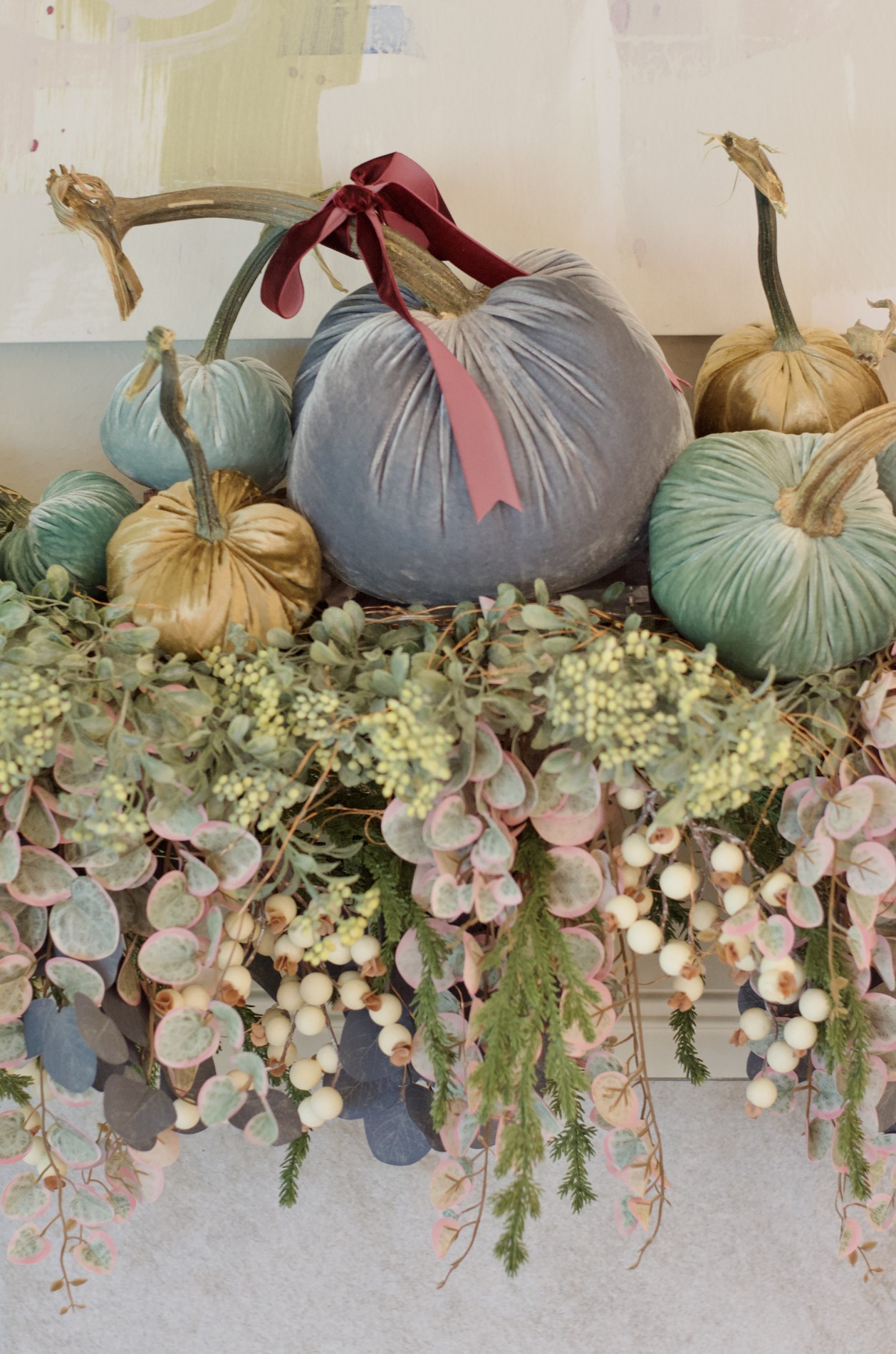 Close-up of blue-gray, green, and gold velvet pumpkins with natural stems, burgundy ribbon, amber candleholders, and cascading foliage on the mantel.